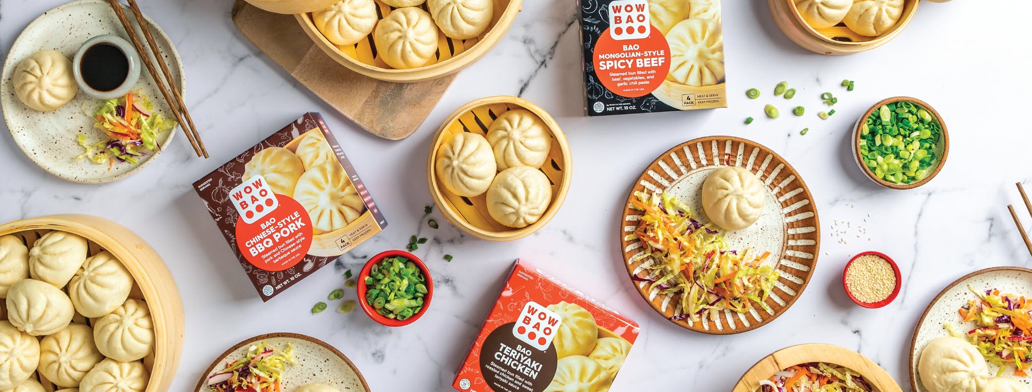 Assorted steamed buns and packets of Wow Bao on a white marble surface, with garnished bowls of green onions, shredded vegetables, and dipping sauce.