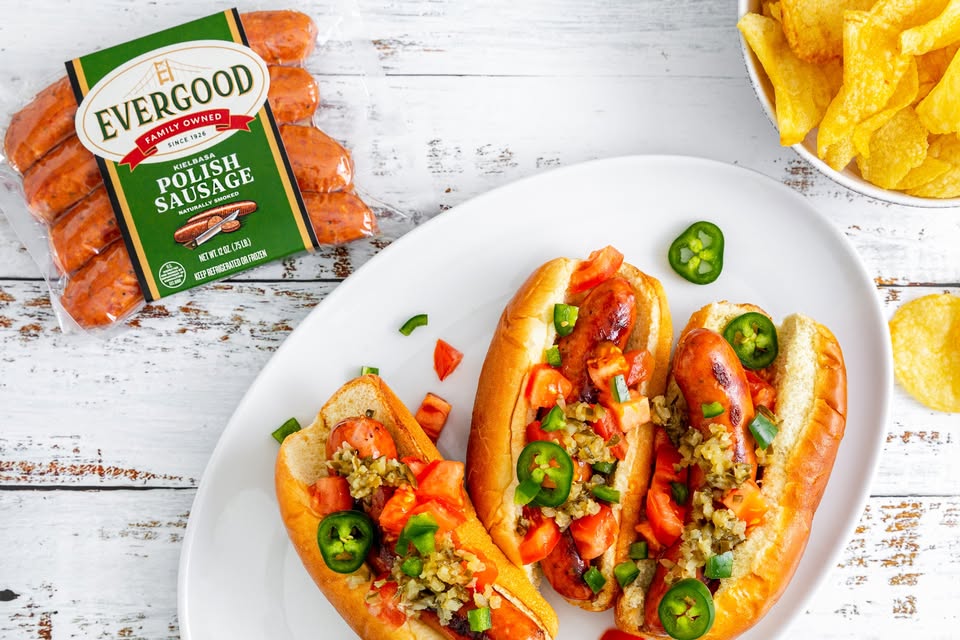 Three sausage hot dogs topped with chopped tomatoes, relish, and sliced jalapeños on a white plate. A pack of Evergood Polish sausage and a bowl of potato chips are nearby.
