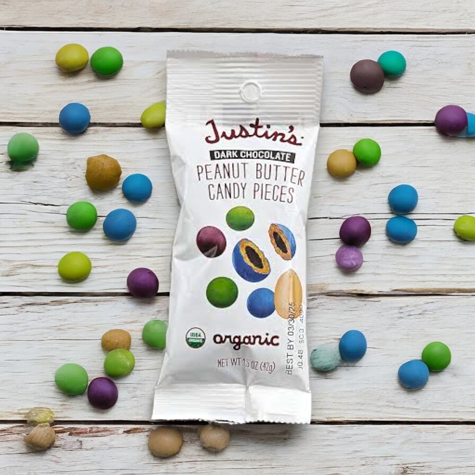 A white packet labeled "Justin's Organic Peanut Butter Candy Pieces-1.5 oz- 1 Count" is surrounded by colorful candy pieces on a wooden surface. The package, featuring images of the candies and an organic label, promises a high-quality indulgence for those craving organic peanut butter candy from Justin's.