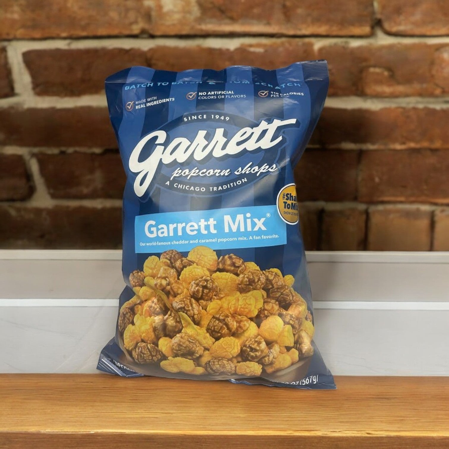 A 20 oz. bag of Garrett Popcorn's Cheddar and Caramel Mix sits on a wooden shelf against a brick wall, highlighting its irresistible blend of cheddar cheese and rich caramel popcorn.