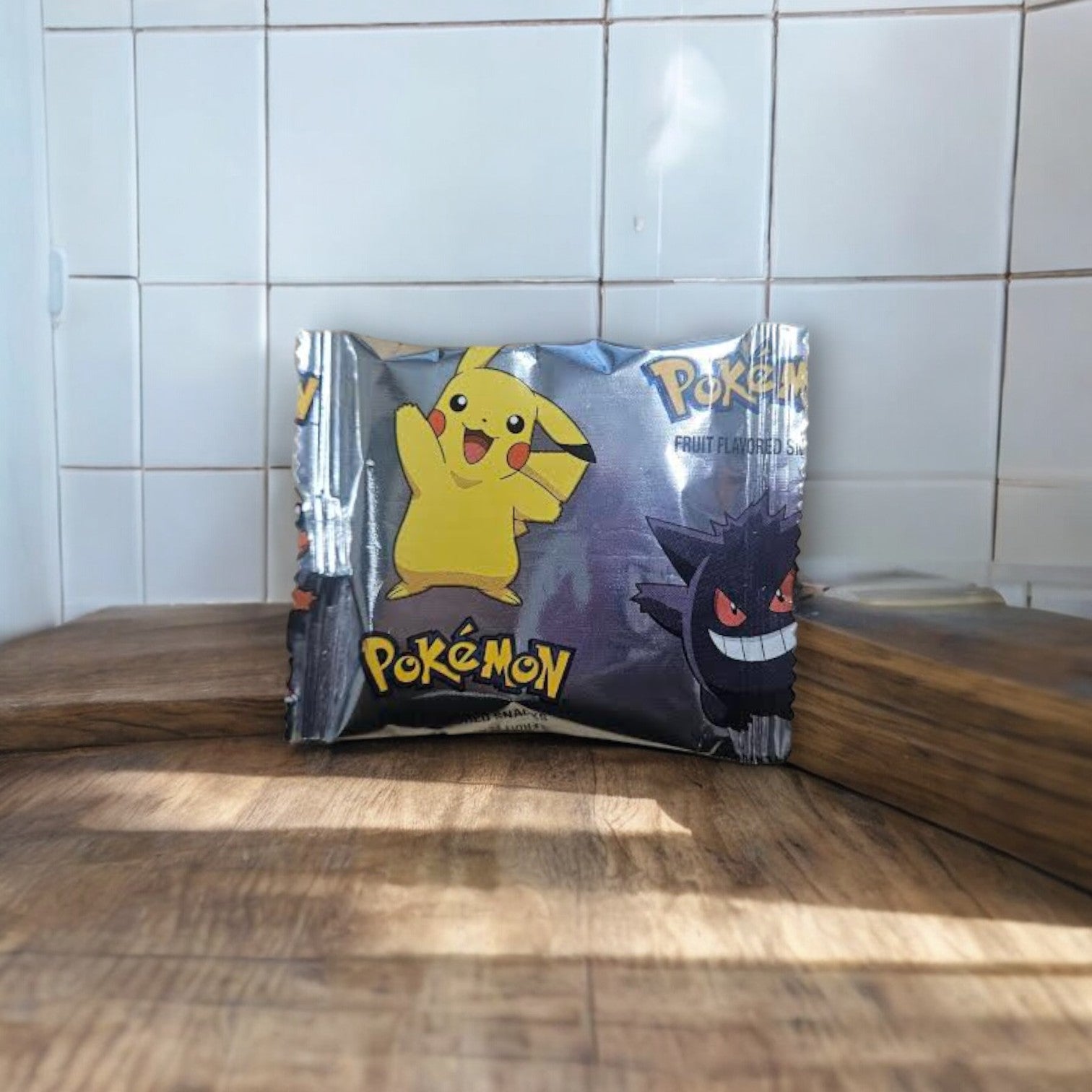 A small, sealed packet of General Mills' Pokémon Gluten Free Fruit Flavored Snack Treat Pouches, featuring cartoon characters Pikachu and Gengar, set against a wooden surface and white tiled wall background.