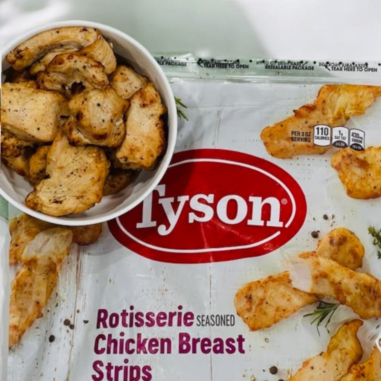 A package of Tyson Rotisserie Seasoned Unbreaded Chicken Breast Strips with a serving in a white bowl placed above the unbreaded chicken.