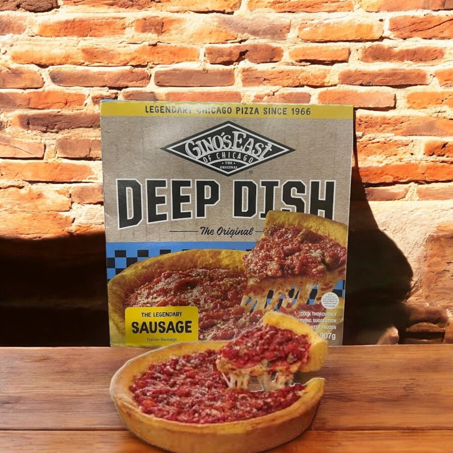 A box labeled "Gino's East Pizza Chicago Deep Dish Sausage Frozen - 32 Oz-1 Count" sits on a wooden table, featuring a slice with crumbled sausage against a rustic brick wall.