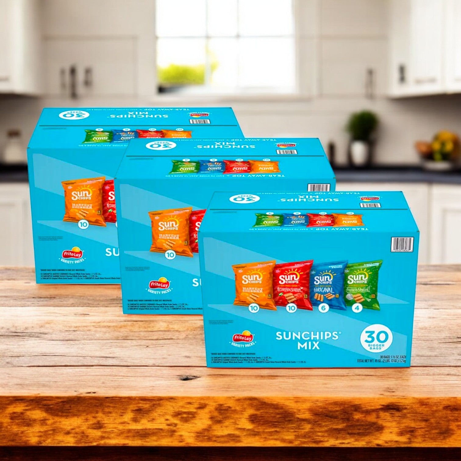 Three Sun Chips Variety Pack boxes, each with 30 single-serve whole grain chip bags, sit on a wooden kitchen countertop. Kitchen cabinets and a window are in the background.