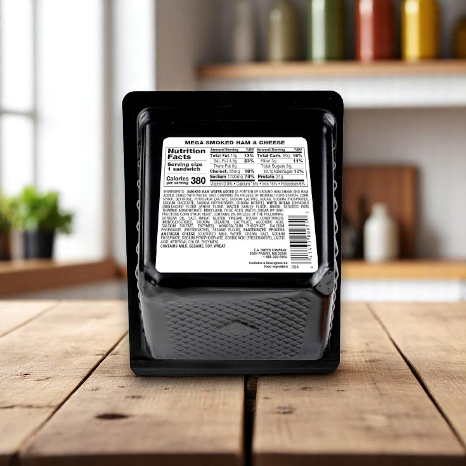A sealed E.A. Sween Market Mega Smoked Ham & Cheese Sandwich Wedge (6.3 oz, individually wrapped) sits on a wooden table, its nutrition label and ingredients visible, with blurred kitchen shelves in the background.