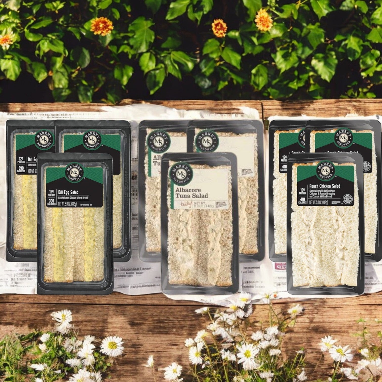 E.A. Sween’s Deli Salad Variety Pack—featuring 3 Chicken Salad Sandwiches, 3 Dill Egg Salads, and 3 Albacore Tuna Salad Sandwiches—is displayed on a wooden surface outdoors with floral and green décor in the background.