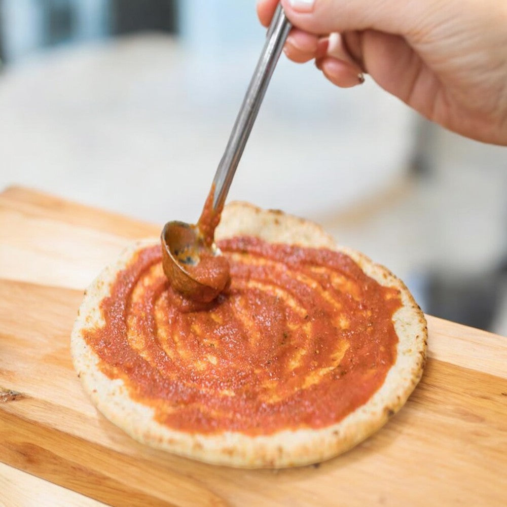 A hand is using a spoon to spread tomato sauce on a Rich's Gluten Free Seasoned Cauliflower Pizza Crust, 10 inch, placed on a wooden surface.