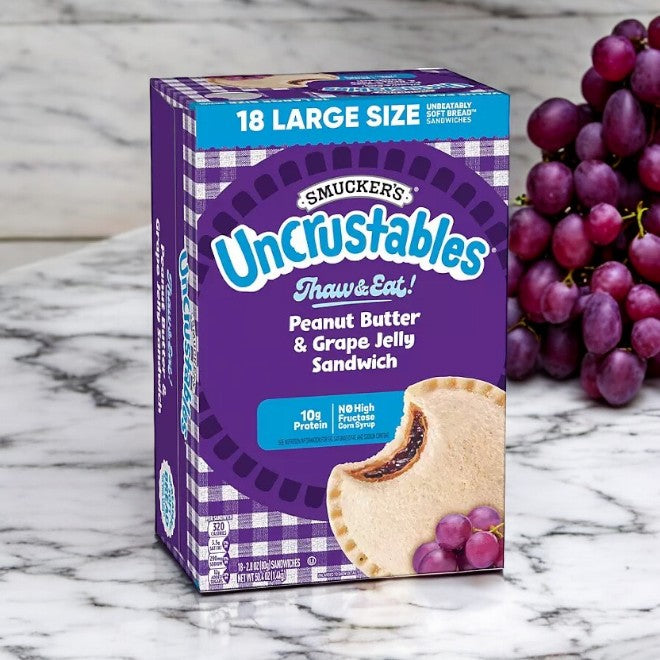 A box of "Uncrustable Smuckers" Grape Frozen Sandwiches, 50.4 oz, 18 Count is displayed on a marble surface. The packaging highlights "18 Large Size" sandwiches with 10g protein per serving and 0g high fructose corn syrup.