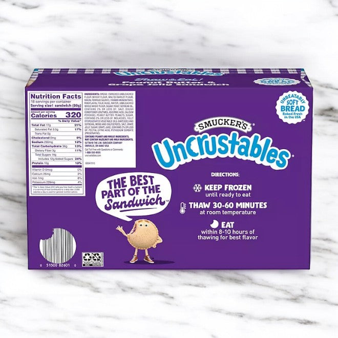 A box of Uncrustable Smuckers Grape Uncrustables Sandwiches, featuring peanut butter and grape jelly flavors, sits on a white marble surface. The 50.4 oz box with 18 sandwiches displays nutritional information, product name, preparation instructions, and ingredients for these delightful frozen treats.