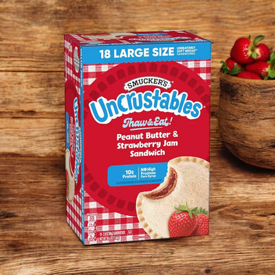 A box of Smucker's Uncrustables Peanut Butter and Strawberry Jam Sandwiches 50.4 oz, 18 Pack, sits on a wooden surface, with a wooden bowl of strawberries in the background. These convenient delights from Uncrustable Smuckers are perfect for any quick snack moment.