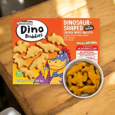 A 67 oz box of Yummy Dino Buddies Chicken Nuggets from the brand Yummy, made from white meat chicken and rich in omega-3 fatty acids, sits next to a white bowl filled with the cooked dinosaur-shaped nuggets on a wooden surface.