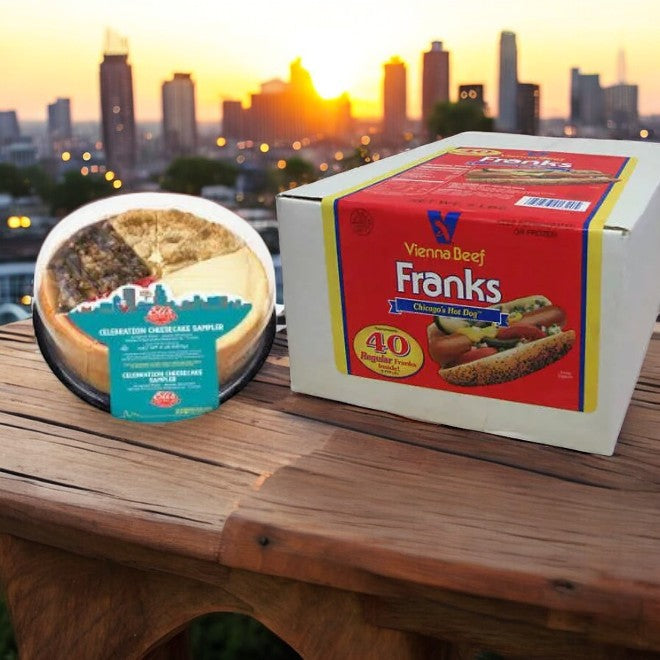 An Elis Cheesecake Sampler and a box of Vienna Beef Franks 40-Pack from Easy Lunches adorn the table, set against the stunning city skyline and vibrant sunset—authentic Chicago staples. Enjoy free shipping!.