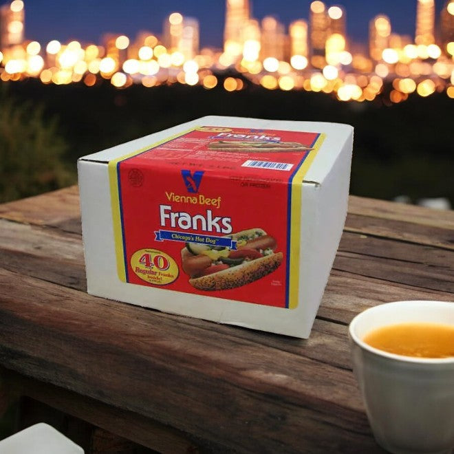 An Elis Cheesecake Sampler & Vienna Beef Franks 40-Pack Hot Dogs from Easy Lunches rests on a wooden surface next to a steaming cup of tea, with Chicago's skyline artfully blurred in the background.