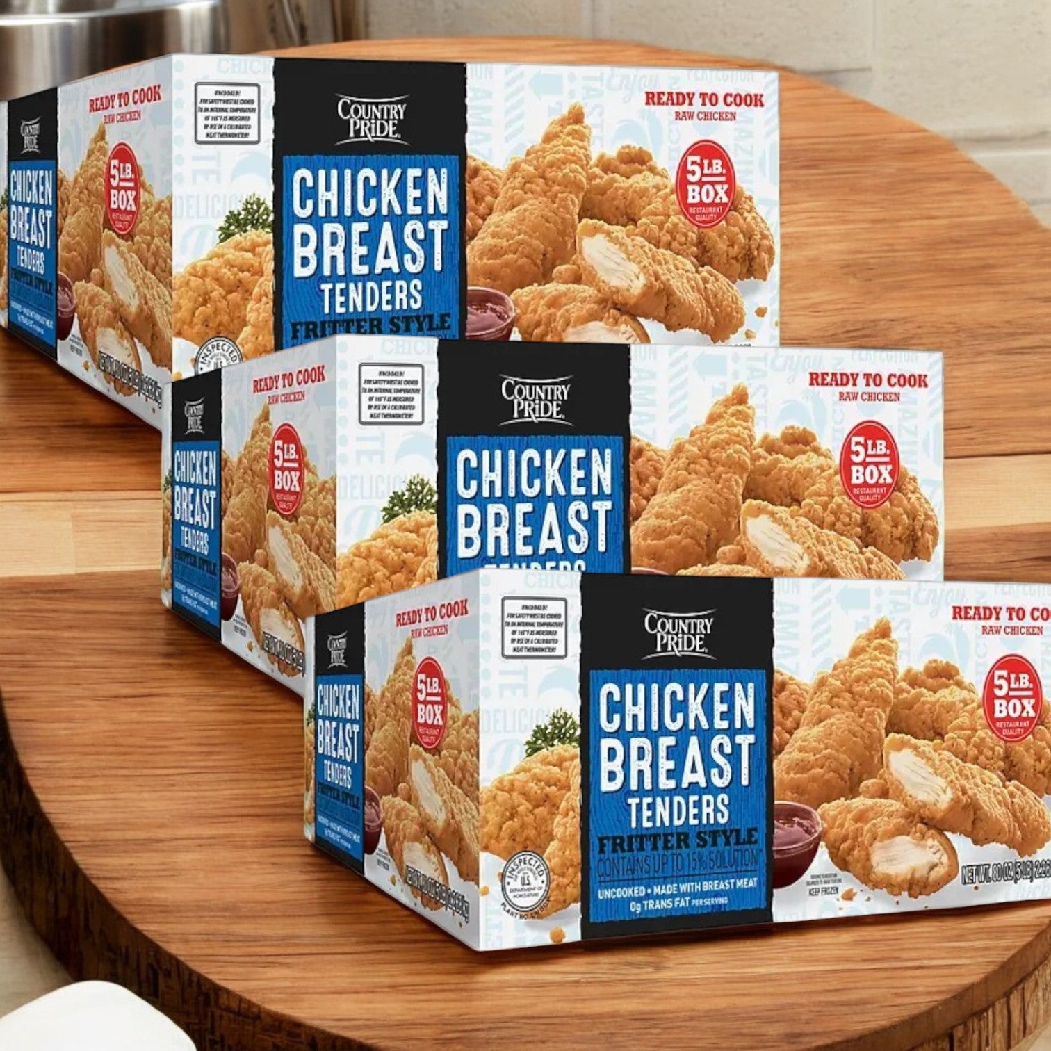 Boxes of Country Pride Ready to Cook Chicken Breast Tenders, each 5 lbs., are arranged on a wooden surface. The packaging features tempting images of fried tenders and promises no preservatives.