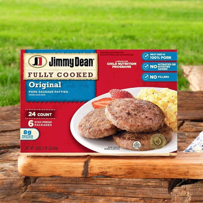 The packaging of the 24-count Jimmy Dean Fully Cooked Original Pork Sausage Patties, presented as a tasty breakfast choice with 8g of protein per serving, is attractively arranged outdoors on a wooden surface.