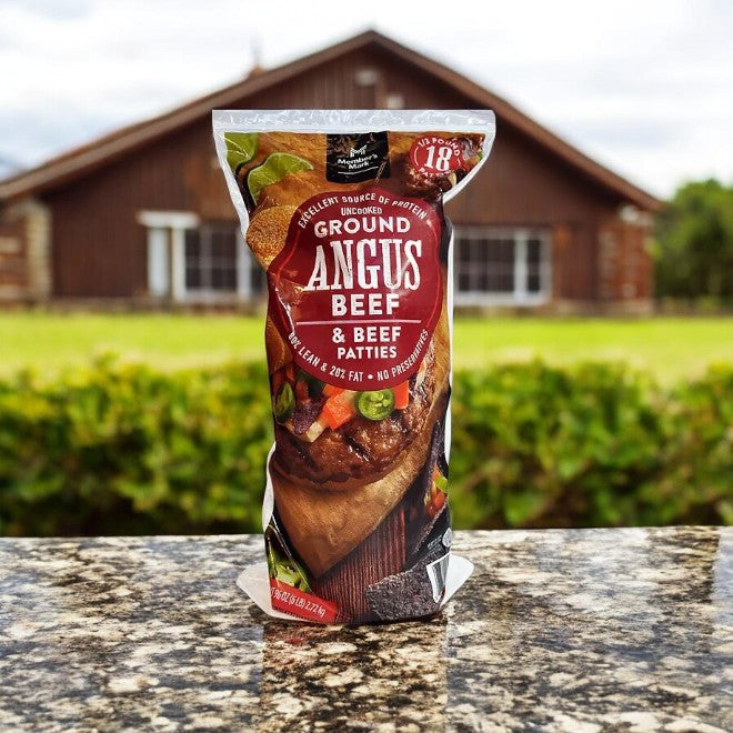 A pack of Member's Mark 80/20 Ground Angus Beef Patties, Frozen, 1/3 lb, is displayed on a stone surface with a blurred wooden house and lush greenery behind.