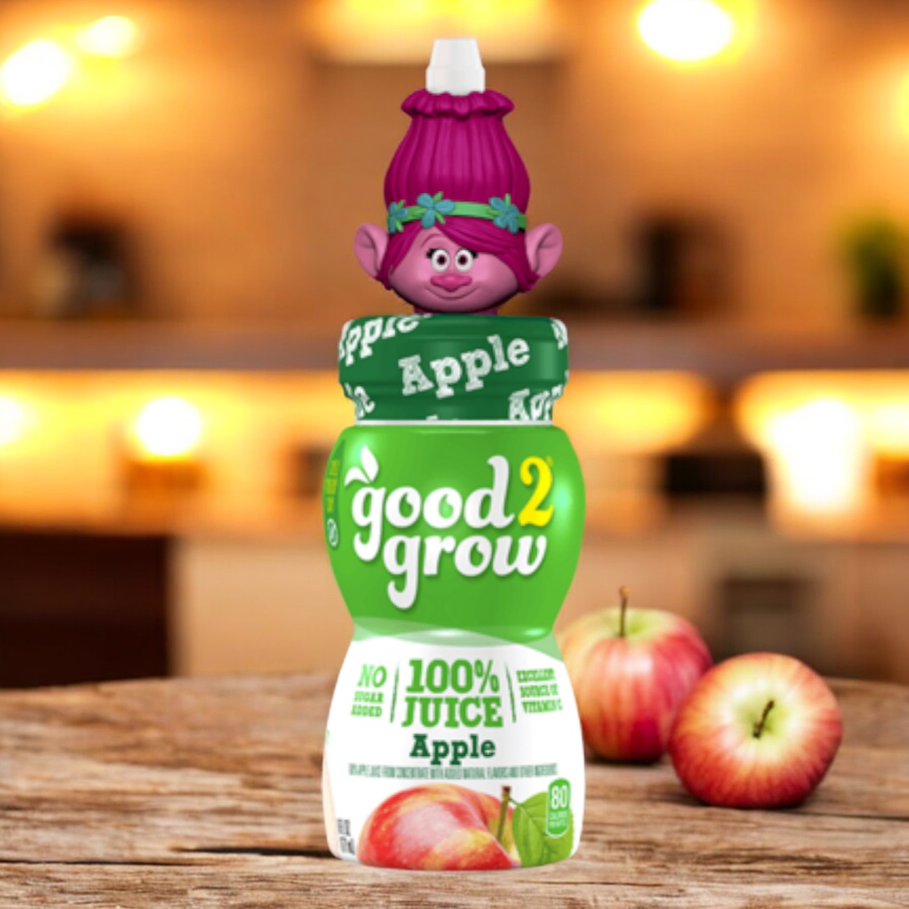 A six-ounce single-serve bottle of Easy Lunches Good2grow Apple Juice, featuring a collectible character top with a purple troll cap, is displayed on a kitchen counter with apples in the background.