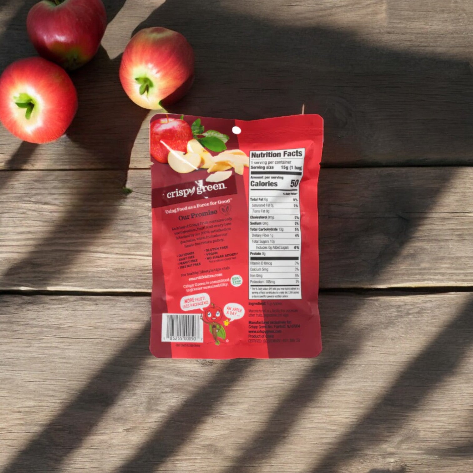 A package of Crispy Fruit All Apple-0.35 oz by Crispy Green lies on a wooden surface next to four whole apples. Sunlight casts shadows over the scene, highlighting these healthy snacks.