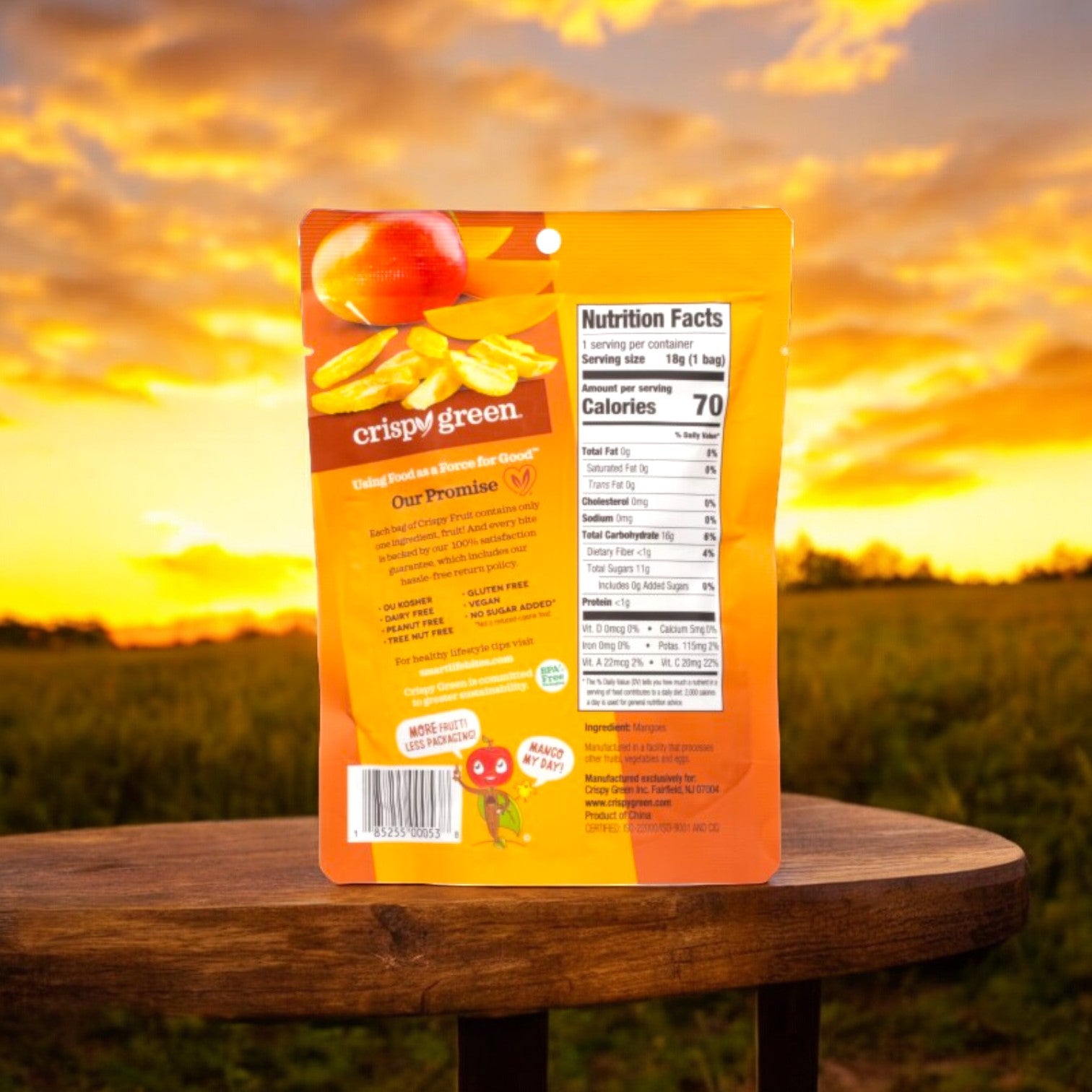 A package of Crispy Green Crispy Fruit All Mango is placed on a wooden surface, with a sunset and field in the background. The back of the package with nutrition facts is visible. Enjoy sustainable snacks like these freeze-dried mango treats, perfect for any occasion.