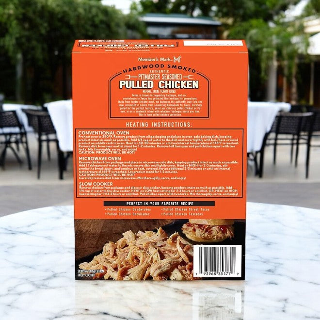 A package of Member's Mark Seasoned Pulled Chicken-1 Count rests on a marble table. The box features cooking instructions and an image of the chicken, with blurred outdoor seating visible in the background.