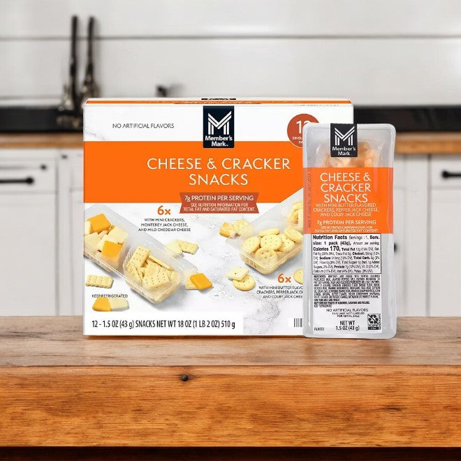 A box of Member's Mark Cheese and Cracker Snacks, 12 Pack-1 Count, rests on a wooden surface, with a softly blurred welcoming kitchen in the background.