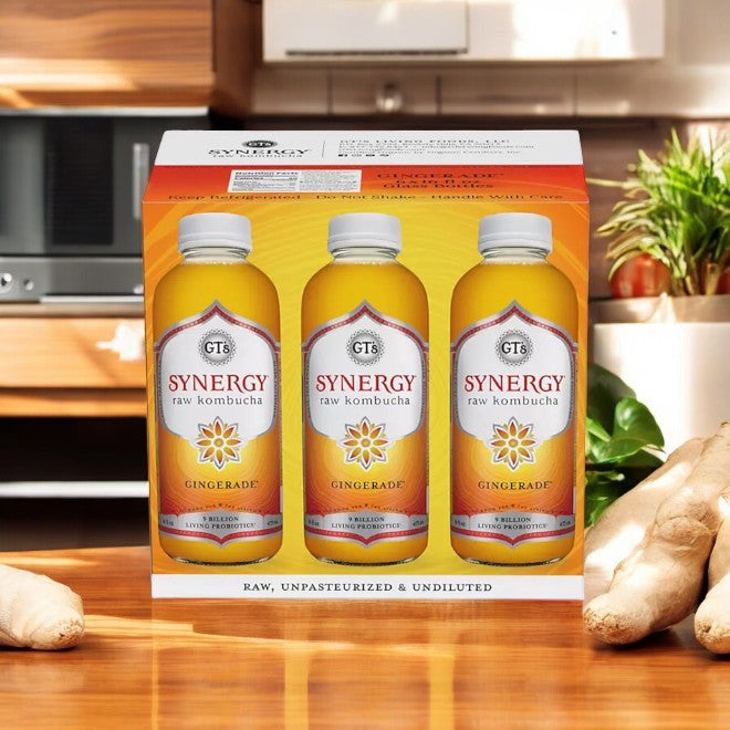 A box of GT's Organic Raw Kombucha Gingerade, featuring six bottles, rests invitingly on a wooden kitchen counter. These probiotic-rich bottles are accompanied by fresh ginger roots and a potted plant in the background.