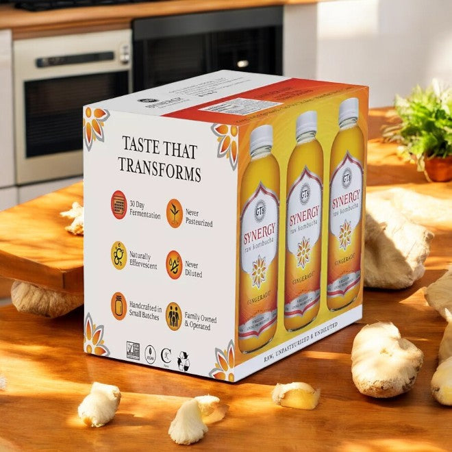 A box of GT's Organic Raw Kombucha Gingerade, 3 Count with a total of 18 bottles, is placed on a wooden kitchen counter, surrounded by fresh ginger pieces, highlighting its probiotic goodness.