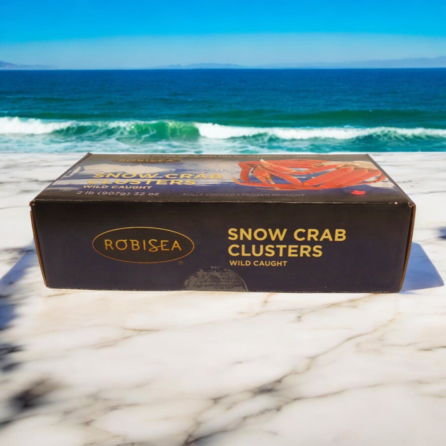 A box of Easy Lunches Robisea Wild Caught Snow Crab Clusters, 2 lb., sits elegantly on a marble surface with an ocean view in the background, sparking thoughts of a decadent Surf & Turf Feast.