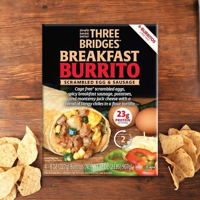 A box of Three Bridges Egg and Sausage Breakfast Burrito, featuring 8 oz burritos in a pack of four, sits on a wooden table surrounded by tortilla chips. This protein-packed breakfast option proudly displays its 23g protein content, showcasing cage-free scrambled eggs as a highlight.