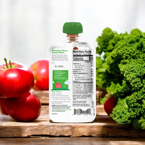 A pouch of Once Upon a Farm Green Kale and Apple organic baby food stands on a wooden surface with kale and apples in the background. Made from an organic fruit and veggie blend, the front shows nutrition facts and instructions for serving this ideal addition to a children's diet.