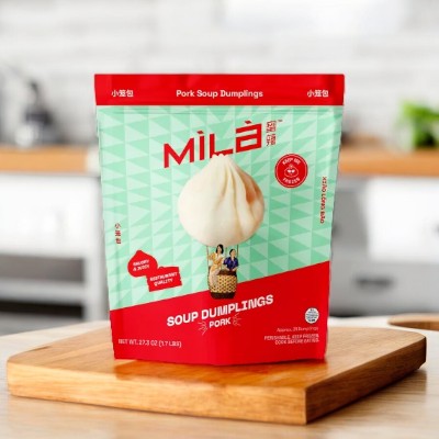 A package of Easy Lunches MiLa Pork Soup Dumplings, Frozen, 31 Count on a wooden board in a kitchen setting. The package is primarily red and green, featuring an image of a dumpling along with product information in both English and Chinese.