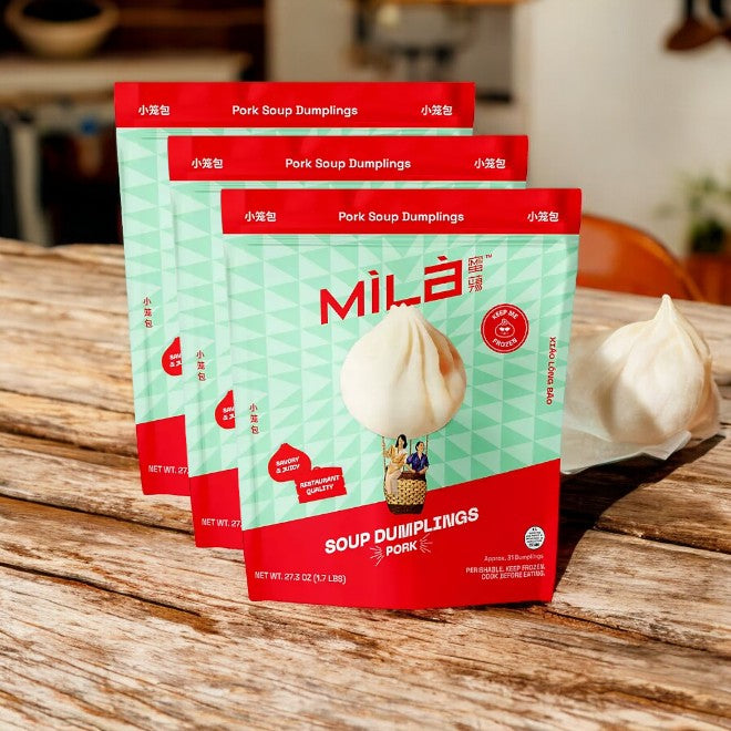 Three packages of 3 - MiLa Pork Soup Dumplings from Easy Lunches are displayed on a wooden surface, with one delicious xiao long bao in front of the packages.