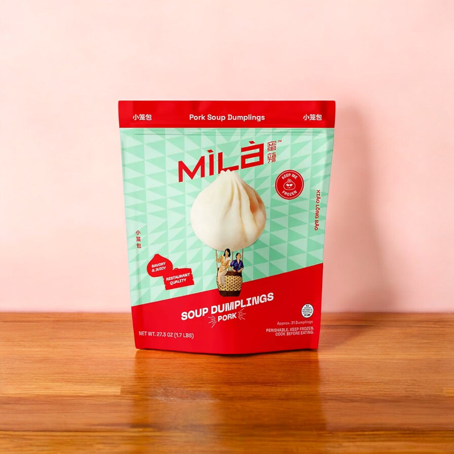 A package of MiLa Pork Soup Dumplings, Frozen, 27.3 oz., sits on a wooden surface against a soft pink background, capturing the tasty allure of xiao long bao.