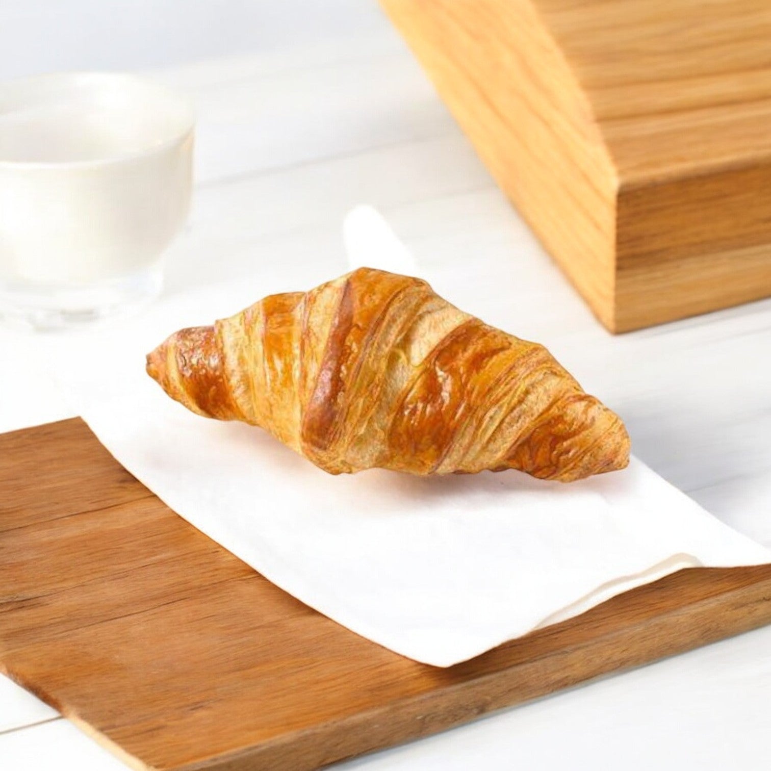 A light and flaky Schar Plain Croissant sits on a white napkin atop a wooden board, with a cup and wooden box in the background. This gluten-free delight from Schar, available in a 7.8 oz package containing ten croissants, is versatile enough for any occasion.