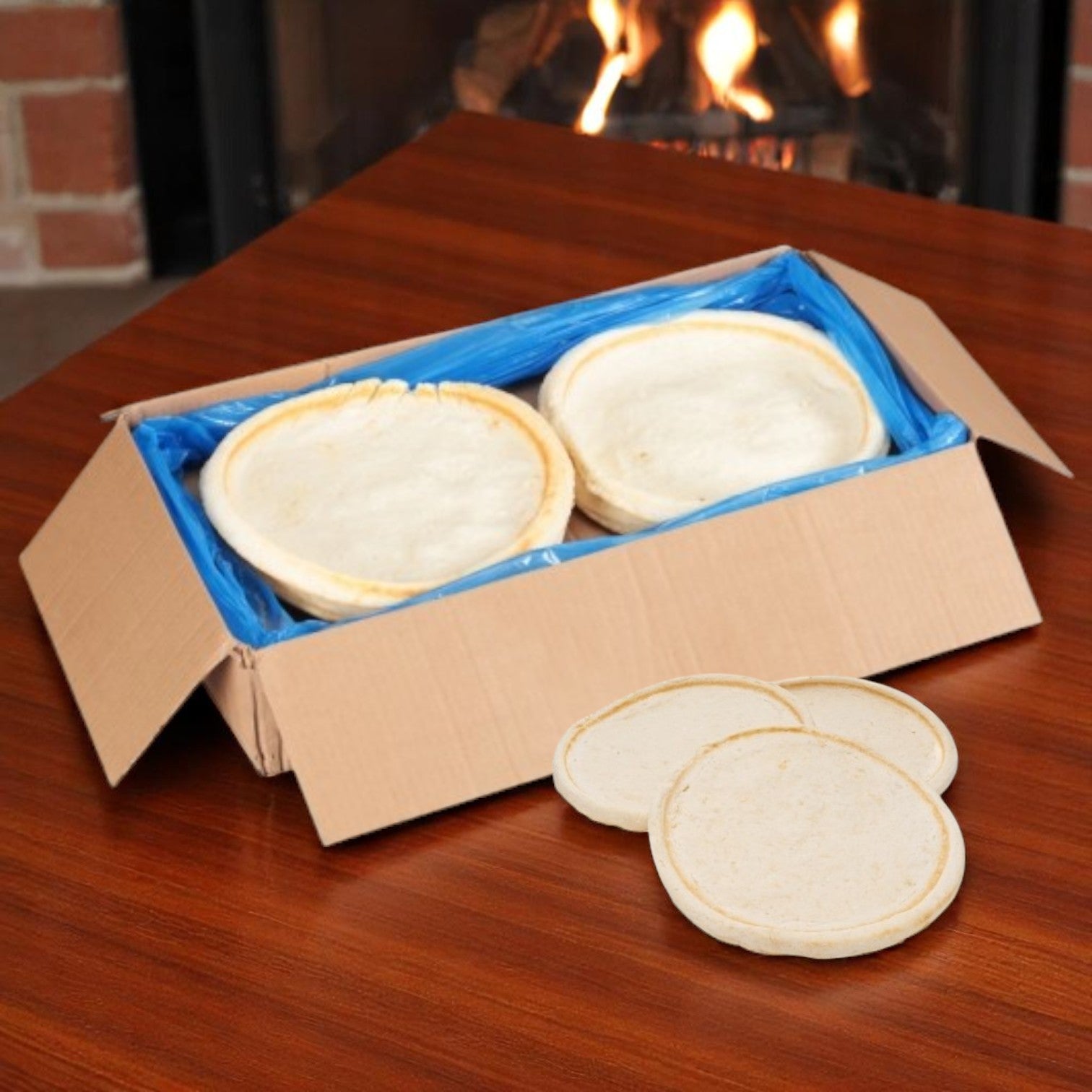 A cardboard box with a blue liner contains Venice Bakery 12" Gluten-Free Pizza Crusts (raised edge, 12.4 oz, 12/case), ideal for foodservice. Three more crusts sit on a table in front of a fireplace.