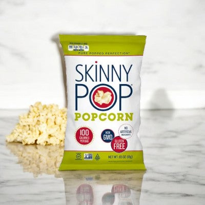 A SkinnyPop Original Popcorn Snack Bag (0.65 oz) is placed on a marble surface, with some scattered whole-grain kernels visible beside it. The packaging highlights that it is 100 calories per bag, non-GMO, and gluten-free, making it a guilt-free snack.