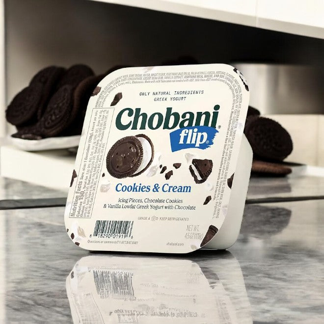 On a marble countertop sits a container from the Chobani Flip Lowfat Greek Yogurt Variety Pack, 4.5 oz, 16 count—temptingly surrounded by chocolate cookies for a delightful treat.