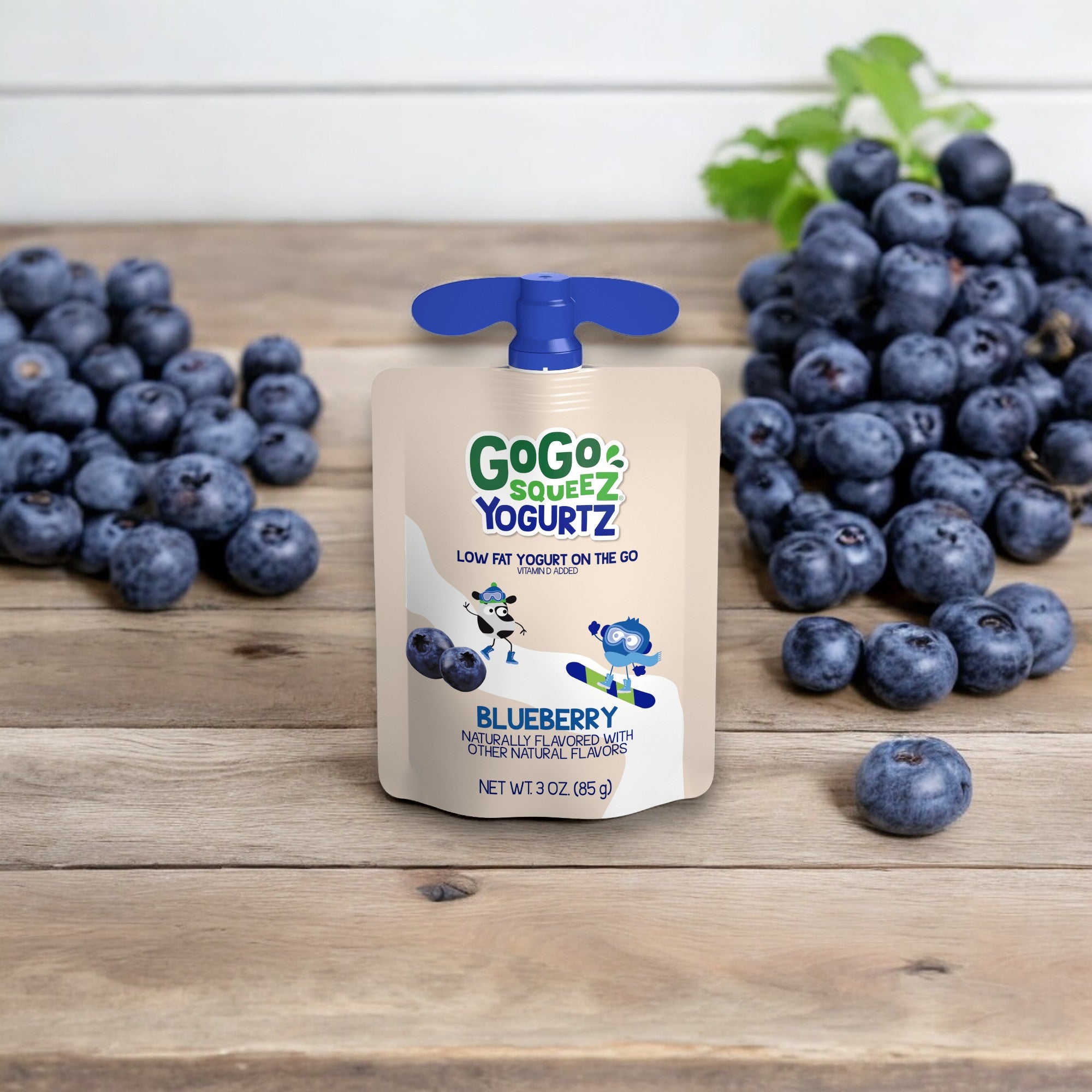 A pouch of GoGo squeeZ® yogurtZ blueberry flavor sits on a wooden surface with fresh blueberries nearby. This 3 oz gluten-free yogurt snack is perfect as an on-the-go treat and promotes itself as a low-fat yogurt option without any preservatives.