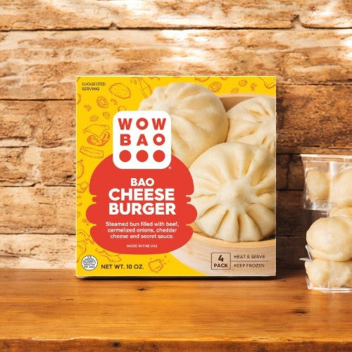 A Wow Bao Cheeseburger Bao-1 Count from Wow Bao rests on a wooden table, highlighting its bold fusion flavors. Nearby, a tempting stack of these delicious buns awaits your taste buds.