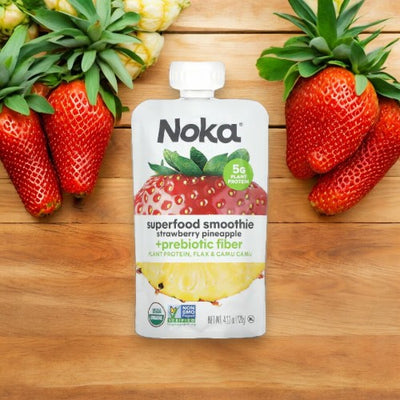 A Noka Organic Strawberry Pineapple Smoothie - 4.22 oz - 1 Count, featuring prebiotic fiber and 5g of plant protein, is surrounded by fresh strawberries on a light wooden surface, making it the perfect on-the-go snack.