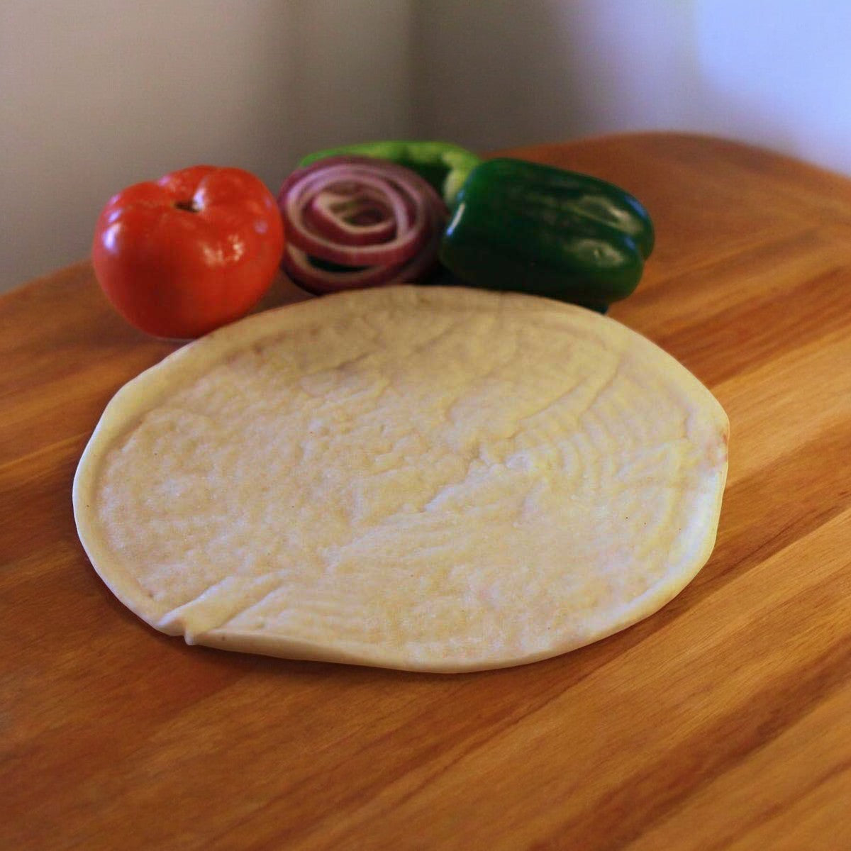 A Venice Bakery Gluten Free Vegan Plain Pizza Crust is positioned on a wooden surface, while vibrant tomatoes, green bell peppers, and sliced red onions enhance the backdrop's color. This vegan-friendly setup offers a deliciously wholesome and inclusive meal option.