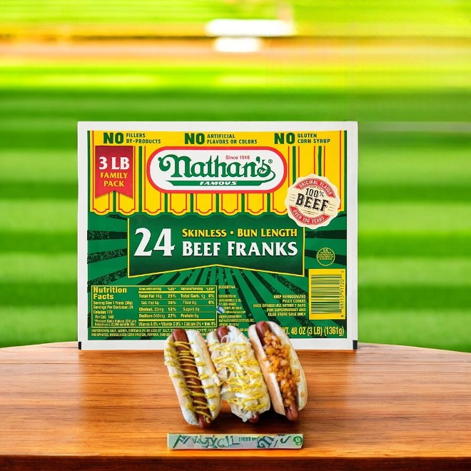 A pack of Nathan's Famous Beef Franks- 1 Count is in a field, with two tasty franks on buns adorned with condiments in the foreground.