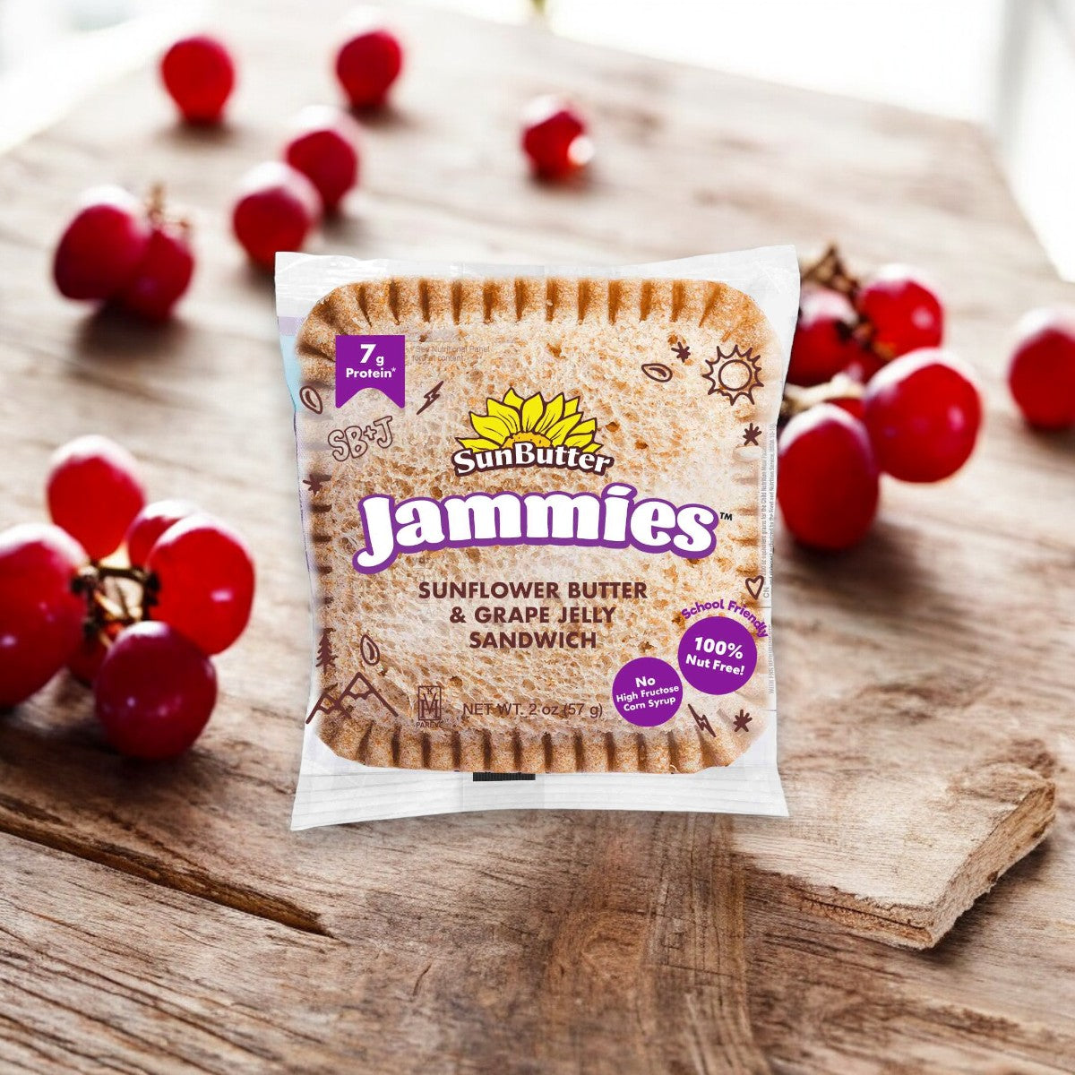 A packaged Sunbutter Jammies Sandwich Sunflower Butter & Grape Jelly 1 Sandwich is placed on a wooden table, with several red grapes in the background, showcasing one of the best nut-free snacks made with whole grains.