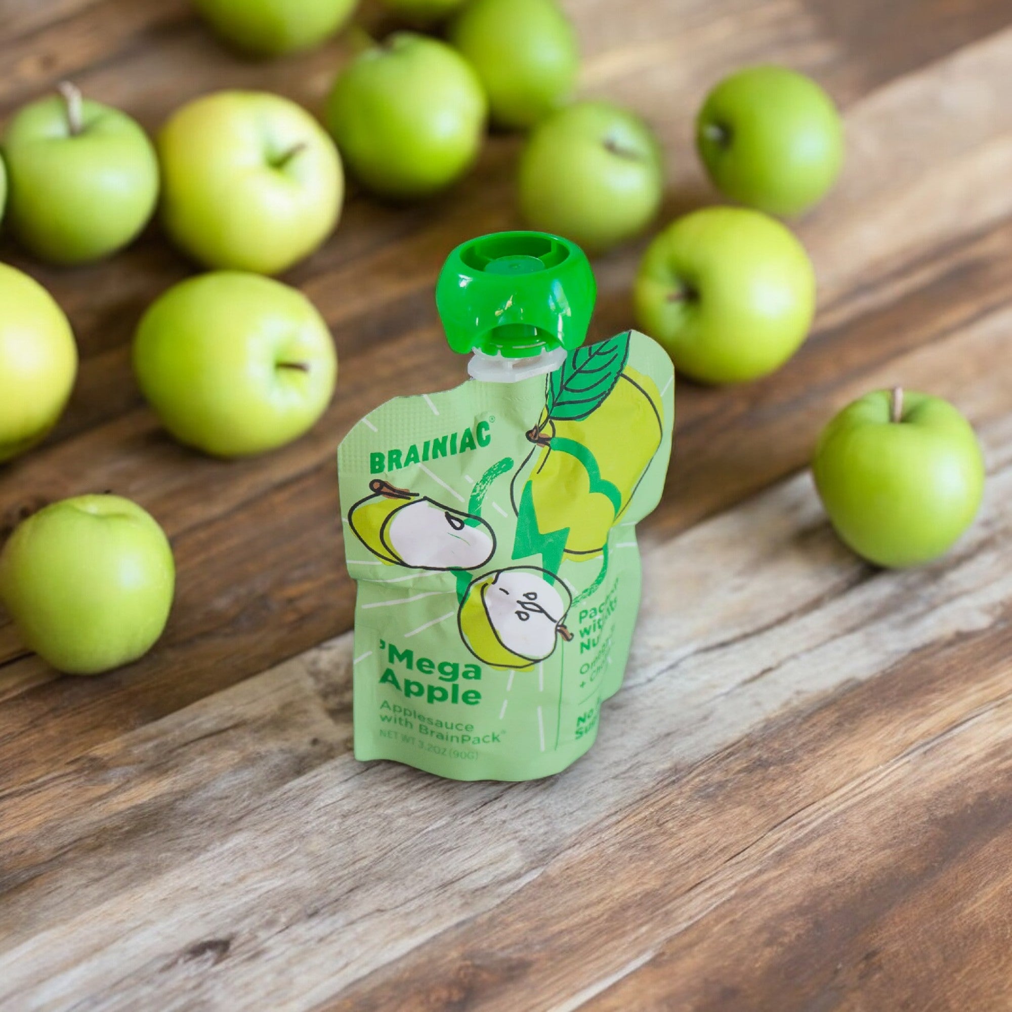 A packet of Brainiac Brain Squeezers Applesauce with Omega-3s, Apple, No Sugar Added (3.2 oz - 1 Count) stands on a wooden surface, with several green apples scattered around in the background. This nutritious snack from Brainiac is fortified with Omega-3s for a brain-boosting treat.