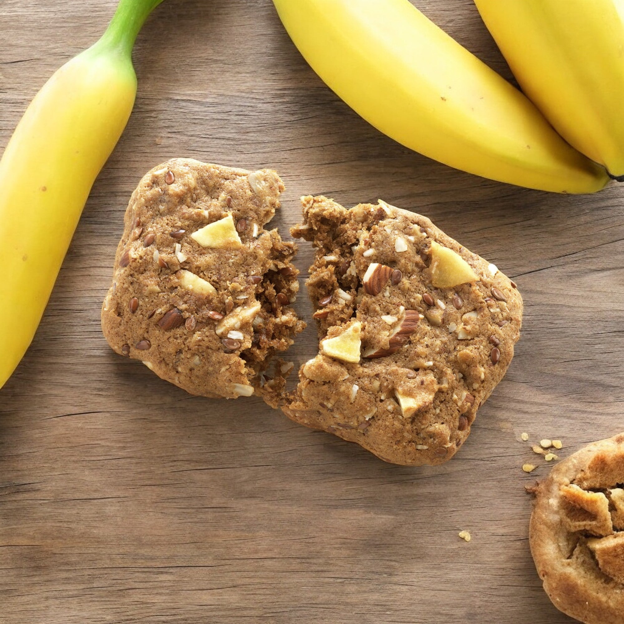 A Simple Mills Soft Baked Almond Flour Bar, Nutty Banana Bread flavor, paleo-friendly and packed with almonds, rests on a wooden surface surrounded by three whole bananas.