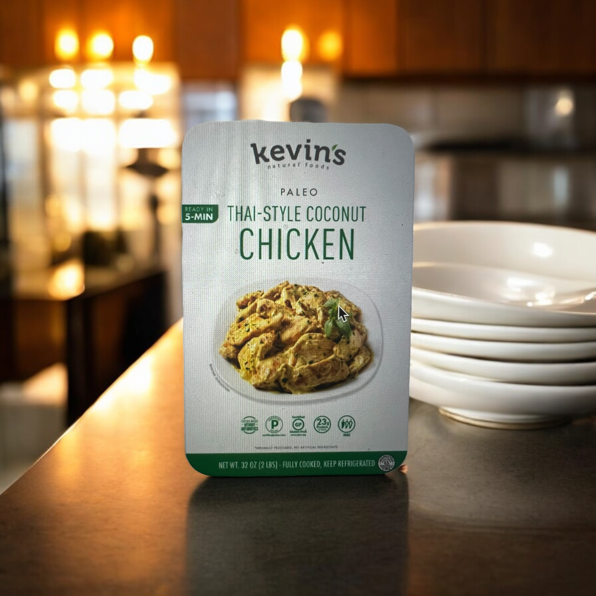 A box of Kevin's Thai-Style Coconut Chicken 1 Box - 16 oz rests on a kitchen counter, accompanied by a stack of white plates in the background. The packaging emphasizes a quick 5-minute preparation time, ensuring an easy meal infused with authentic Thai spices and a rich coconut sauce.