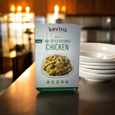 A box of Kevin's Thai-Style Coconut Chicken 1 Box - 16 oz rests on a kitchen counter, accompanied by a stack of white plates in the background. The packaging emphasizes a quick 5-minute preparation time, ensuring an easy meal infused with authentic Thai spices and a rich coconut sauce.