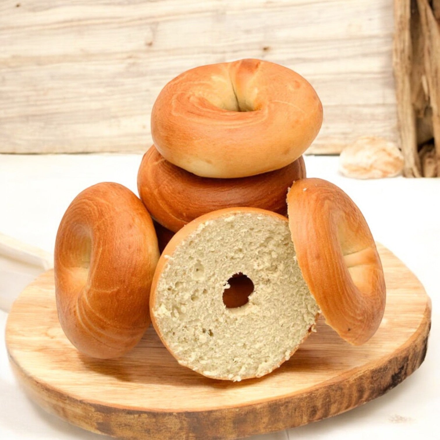 On a wooden board against a light background, there is a stack of Schar Gluten Free Plain Bagels from the 14.1 oz. pack, with one bagel sliced open.