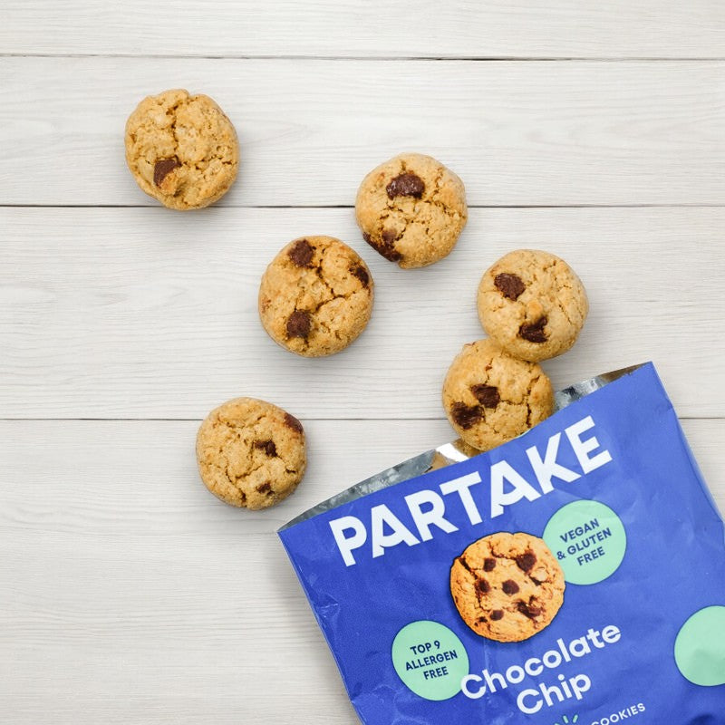 A blue bag labeled "Partake Crunchy Mini Chocolate Chip Cookie Snack Packs, 1 Count" with several crunchy chocolate chip cookies spilling out onto a wooden surface. The bag indicates that the snacks are vegan and allergy-friendly, top 9 allergen-free, and gluten-free.