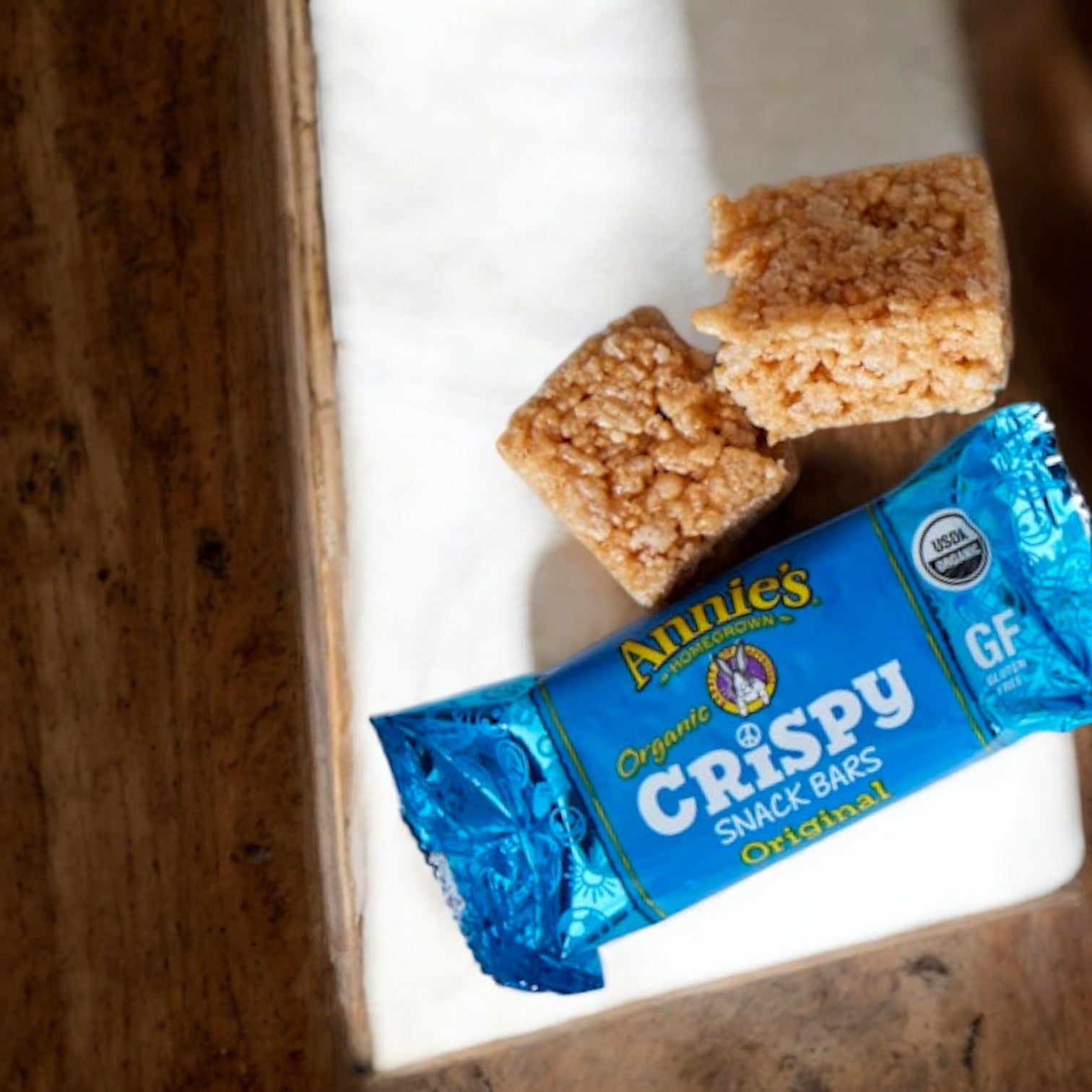 Two Annie's Organic Original Crispy Snack Bars, featuring quinoa crisps and labeled gluten-free, are displayed on a white surface with a wooden background. The blue packaging reads "Annie's Organic Original Crispy Snack Bars.